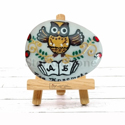 Ръчно рисуван камък с бухал с разперени крила и буквите „А“ и „Б“, поставен върху дървена етажерка.