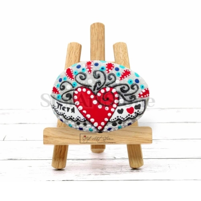 Ръчно рисуван морски камък с изобразено червено сърце и персонализирано име, поставен върху дървена повърхност.